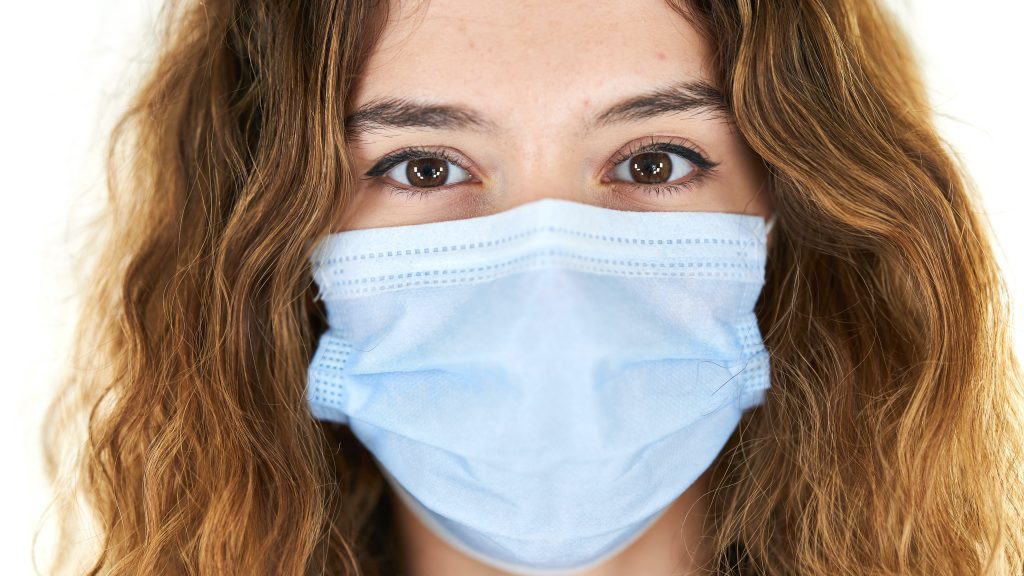 Young girl with alive eyes and brown wavy hair wearing a blue surgical mask