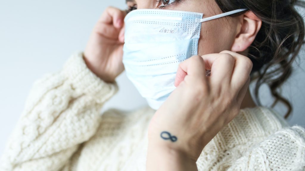 Young adult wearing blue surgical mask and fixing it on her face, with small infinity tattoo on wrist